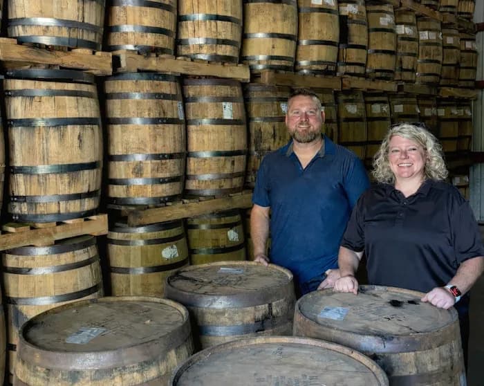 Two people standing in front of a rickhouse wall stacked with barrels